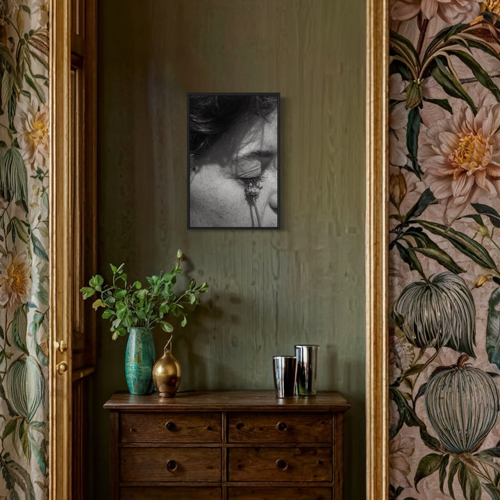 The artwork 'Communion II' by Eunice Pais showcases a black and white photograph displayed in an elegant interior setting. The photograph hangs on a sage green wall, complemented by floral wallpaper featuring peach dahlias and botanical motifs. Below sits a vintage wooden dresser adorned with a turquoise vase holding fresh greenery and metallic decorative objects. Photography artwork by Eunice Pais.