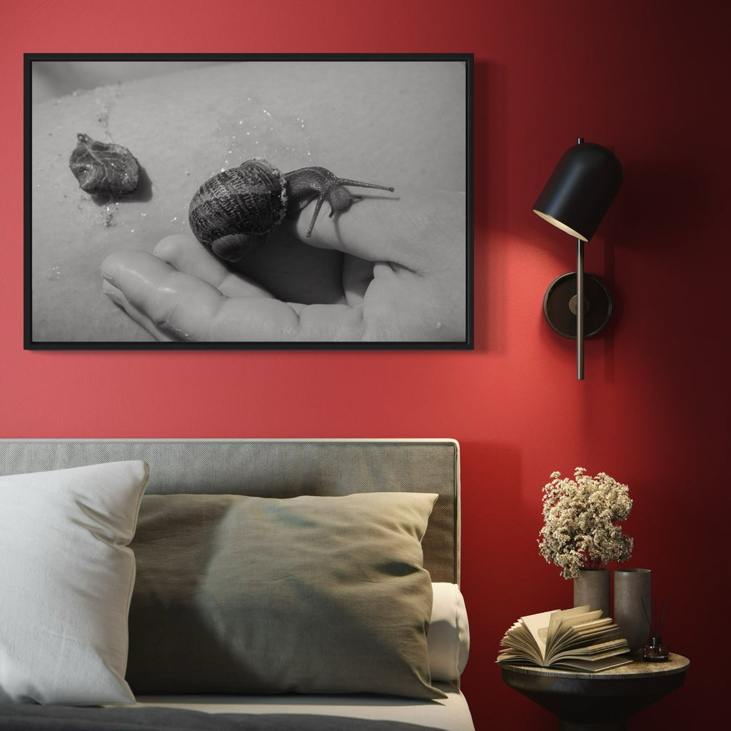 The artwork 'Communion IV' by Eunice Pais shows a striking black and white photograph of a snail crawling across an outstretched palm, creating an intimate dialogue between human and nature. The monochromatic composition emphasizes texture and form, with the snail's spiral shell casting subtle shadows. The minimalist framed photograph hangs on a bold red wall. Photo print by Eunice Pais.