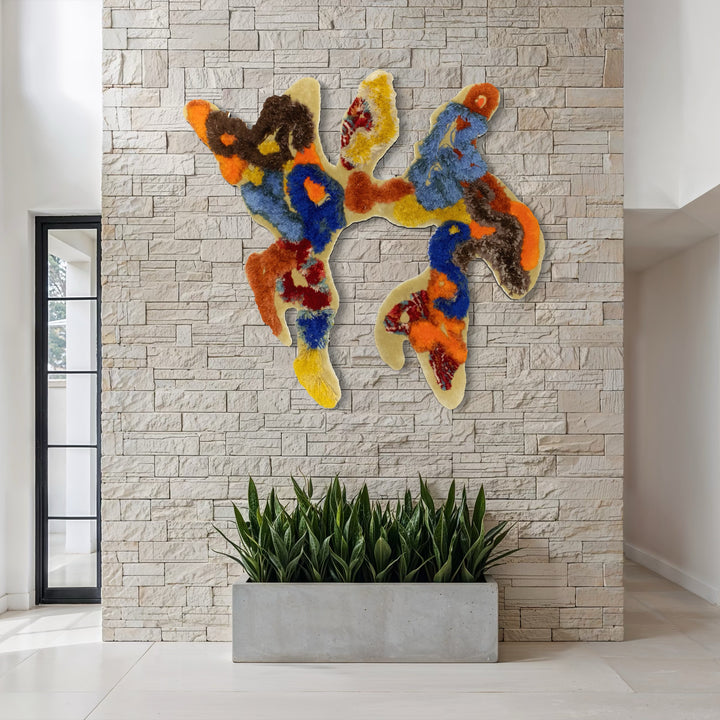 The Crested Ibis Lovers by Abigail Henthorne showcases a vibrant, textured abstract shape in energetic hues of orange, blue, yellow, and brown, mounted on a light stone wall above a minimalist planter with green foliage, creating a dynamic contrast between natural elements and colorful contemporary art.
