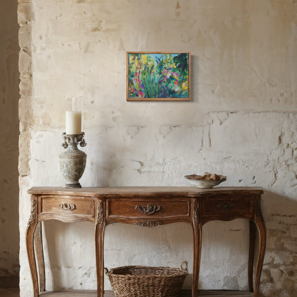 The artwork 'Daisy Horizon' by Maureen Shea features vibrant impressionistic florals with pink daisies and green foliage against a textured turquoise background, oil painted on wood panel, displayed above an antique console table in a rustic setting with whitewashed stone walls. 12x16 inches, framed in warm wood.