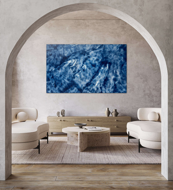 The artwork 'Entangled' by Gina Iannucci features dynamic swirls of deep and light blue spray paint on canvas, creating a mesmerizing abstract pattern reminiscent of ocean waves or stormy skies. The fluid, ethereal composition showcases a rich interplay of textures and tonal variations, displayed prominently on a 96-inch wide canvas against a neutral wall by Gina Iannucci.