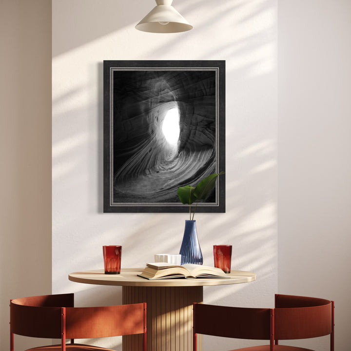 Emergence by Kyle Murphy reveals a luminous natural opening within a smoothly sculpted rock formation, captured in striking black and white. The artwork invites contemplation of light piercing darkness, with delicate textures that evoke depth and quiet intensity, enhancing the serene, minimalist dining space where it hangs.