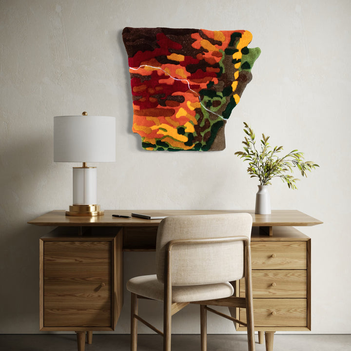 The artwork Endangered Natural State - Fall by Abigail Henthorne features a textured, abstract representation in rich autumn hues of red, orange, yellow, brown, and green. It hangs on a neutral wall above a wooden desk with a beige chair, white lamp, and a minimalist vase holding green foliage, blending natural warmth with artistic expression.