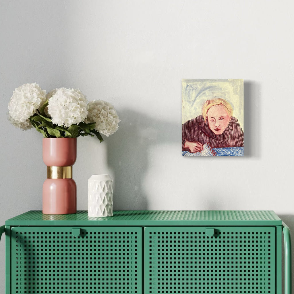 The artwork 'Eve Doing Homework After Eating an Apple from the Garden' by Monica Carrier depicts a contemplative figure leaning over a blue surface against a swirling yellow background. Oil on linen painting showcases expressive brushstrokes in burgundy and pastel tones. Displayed above a green perforated cabinet with white hydrangeas in a pink and gold vase. 10x8 inches.