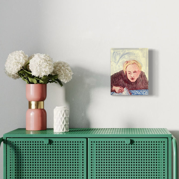 The artwork 'Eve Doing Homework After Eating an Apple from the Garden' by Monica Carrier depicts a contemplative figure leaning over a blue surface against a swirling yellow background. Oil on linen painting showcases expressive brushstrokes in burgundy and pastel tones. Displayed above a green perforated cabinet with white hydrangeas in a pink and gold vase. 10x8 inches.