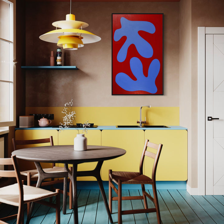 The artwork 'FERN II' by Shadia Sabagh features bold organic shapes in vibrant blue against a striking red background, displayed in a contemporary kitchen setting with yellow cabinetry, modern pendant lighting, and wooden dining furniture. The minimalist abstract composition presents a 48x30 inch acrylic painting that evokes the natural curves and forms of fern fronds.