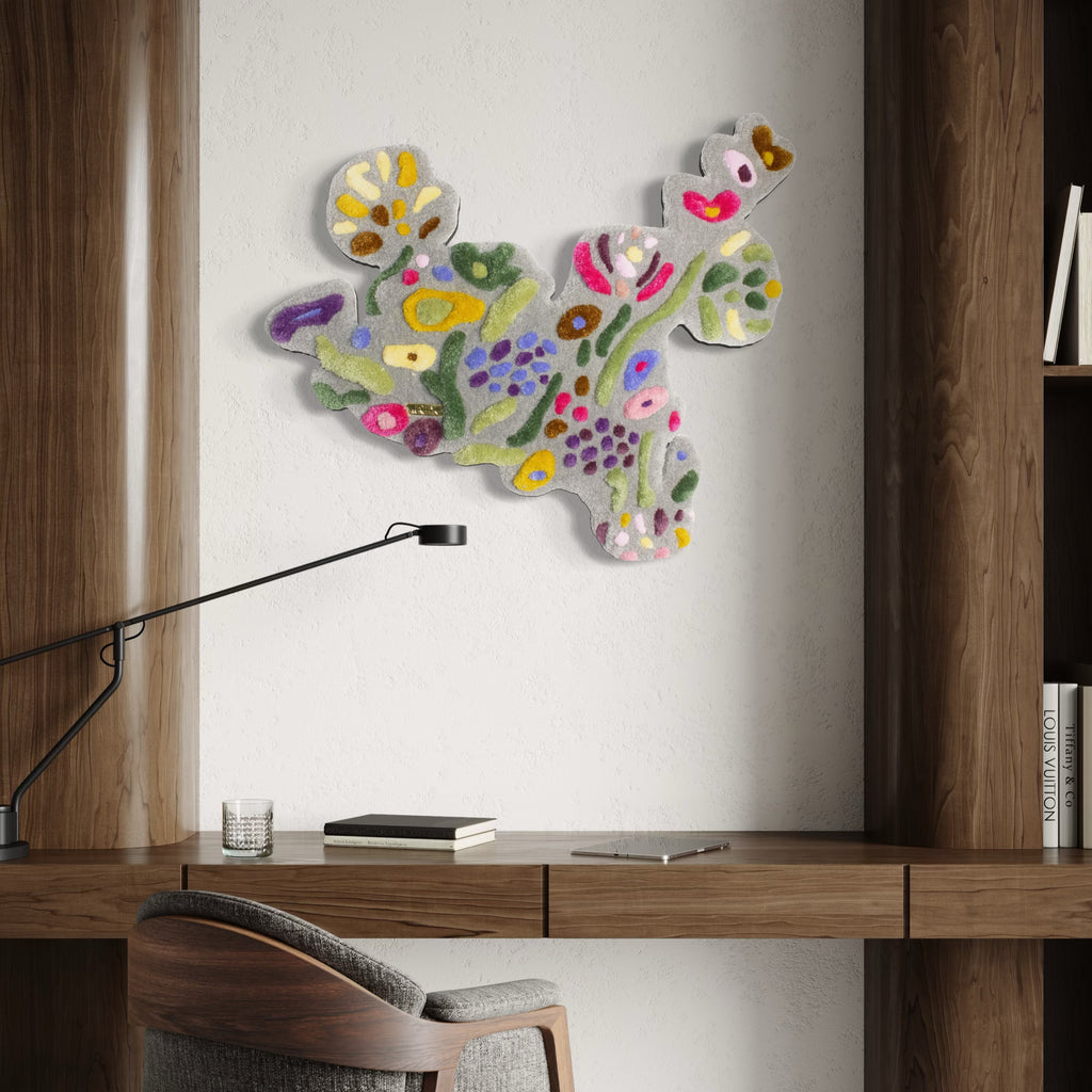 The Flower Arrangement by Abigail Henthorne presents an abstract, textured wall piece with soft pastel hues of pink, green, yellow, purple, and white, evoking a lively mix of organic, floral-inspired shapes that add a gentle yet energetic touch to a modern workspace with warm wooden accents.