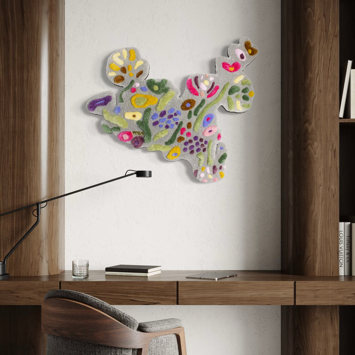 The Flower Arrangement by Abigail Henthorne presents an abstract, textured wall piece with soft pastel hues of pink, green, yellow, purple, and white, evoking a lively mix of organic, floral-inspired shapes that add a gentle yet energetic touch to a modern workspace with warm wooden accents.
