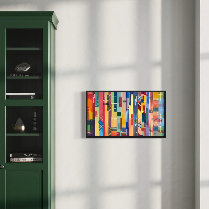 The artwork 'Fragmented Legacy' features a vibrant abstract watercolor composition of vertical geometric shapes in bold primary colors. The painting displays intersecting rectangles and linear patterns in red, blue, yellow, and orange, creating a dynamic urban-inspired mosaic effect within a black frame. Shown mounted on a white wall beside a forest green bookshelf. By Arthur Norcome.