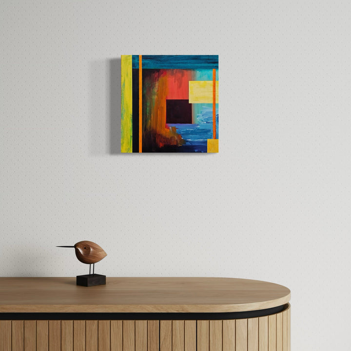 The artwork 'Gentle Rain' by Vanessa Ryan features an abstract geometric composition with vibrant color blocks including turquoise blue, sunset orange, and bright yellow. A striking black square anchors the center, while bold vertical lines create dynamic movement across the 12x12 acrylic canvas, evoking a modern, contemporary aesthetic with rich textural depth.