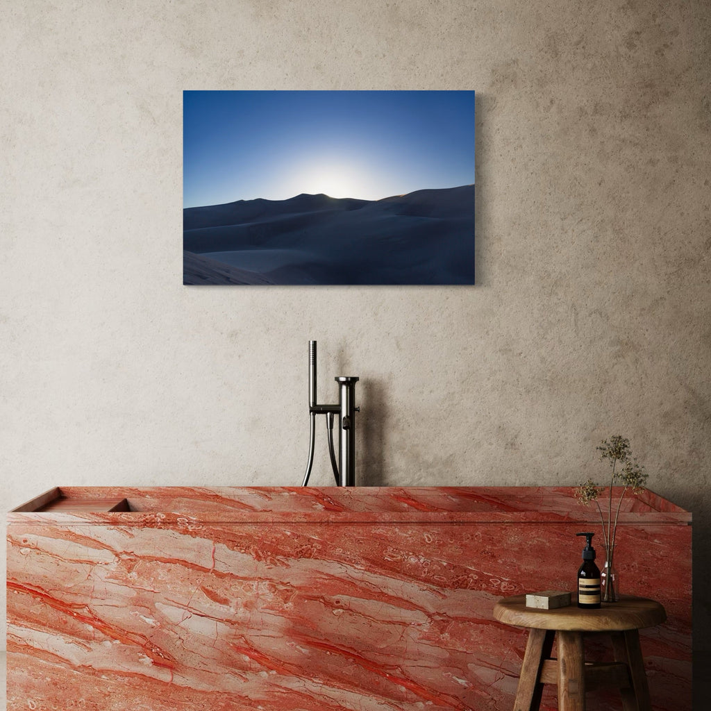 The artwork 'Hiding Sun' by Cavier Coleman captures a serene landscape photograph featuring silhouetted mountain ridges against a brilliant blue sky. The sun rests just behind the mountain peak, creating a luminous glow effect. The composition showcases dramatic contrast between dark mountain shapes and bright sky, photographed in a minimalist style at 24x36 inches by Cavier Coleman.
