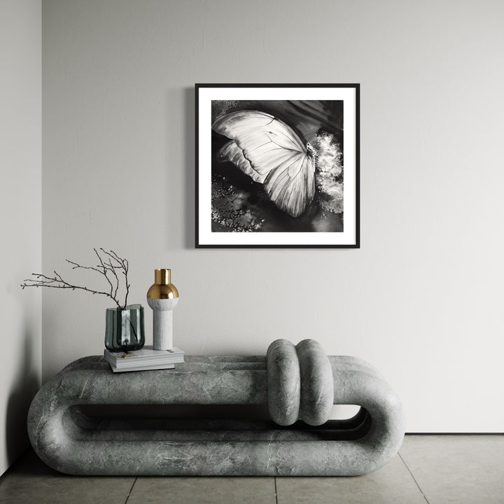 The artwork 'Iridescence' by Cat Lencke features a striking black and white watercolor study of a luminous butterfly wing. The delicate details of the wing structure are captured in dramatic contrast, with soft textural elements and ethereal light effects creating a mysterious atmosphere. The image is presented in an elegant black frame against a white background.