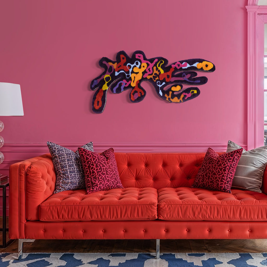 The King Vulture by Abigail Henthorne is an abstract, textured wall art piece featuring bold, organic shapes in vibrant shades of orange, red, purple, and yellow, mounted on a rich pink wall above a tufted coral sofa with patterned cushions, creating a striking focal point with a dynamic yet harmonious blend of color and form.