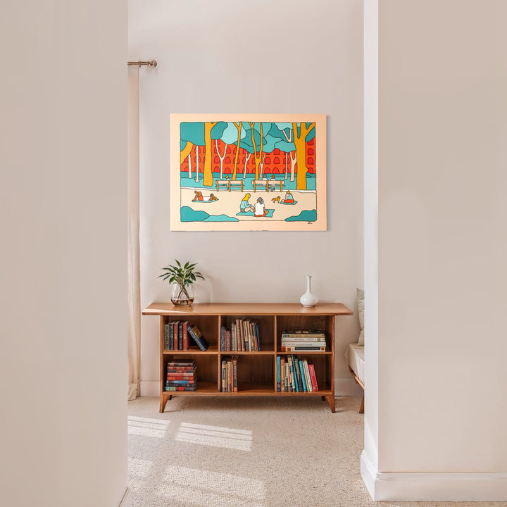 NYC Vibes by Alimo features a colorful scene of people enjoying a sunny urban park framed by tall trees and distinctive red buildings, displayed above a wooden bookshelf in a bright, minimalist room, capturing a playful and lively artistic atmosphere.