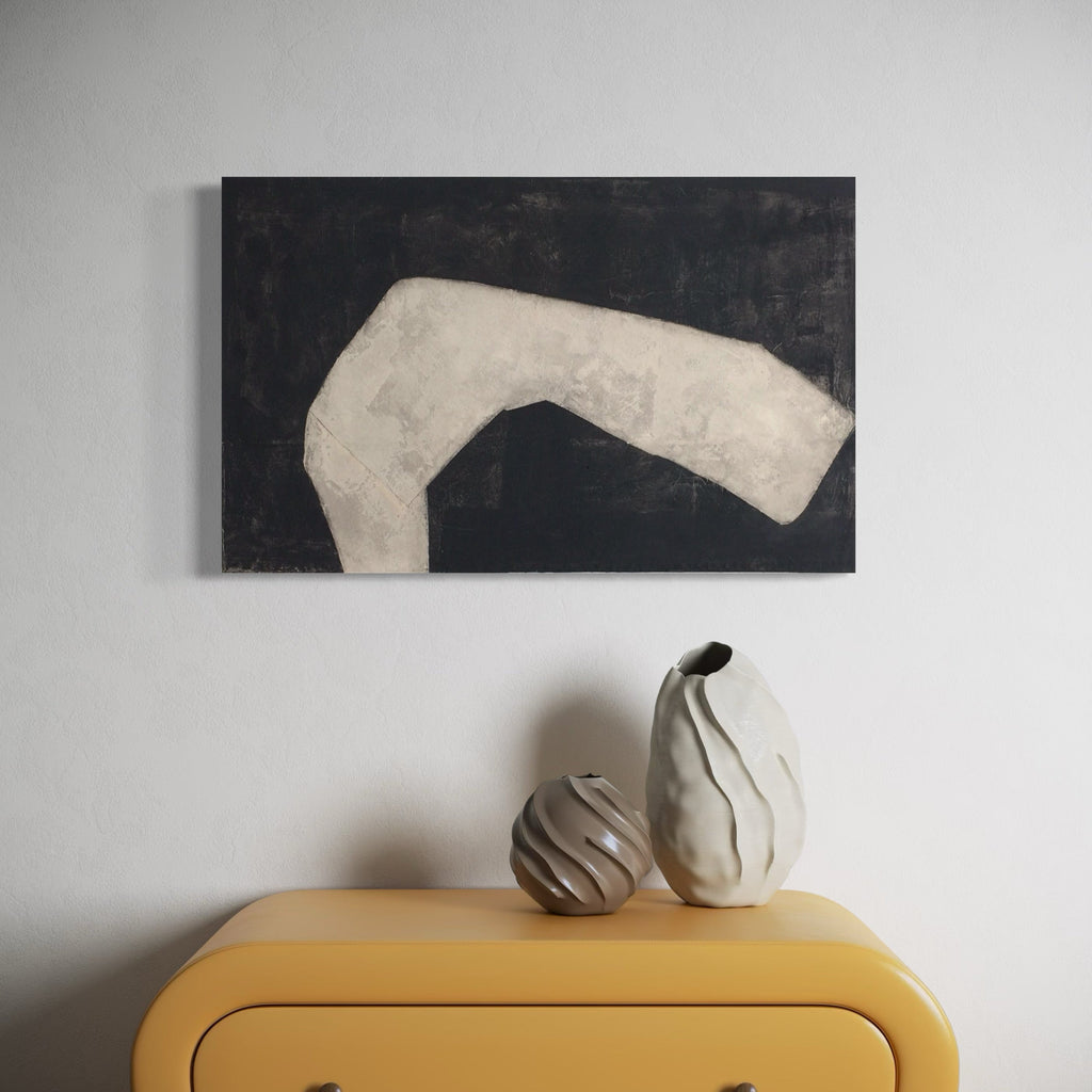 The artwork 'Pale Form on Black' by Diane Englander features a striking minimalist composition with an abstract white curved shape against a deep black background. The textural mixed-media piece combines paper, fabric, and acrylic on canvas, creating a bold contrast and sculptural presence. Displayed above modern decorative vessels on a yellow cabinet.
