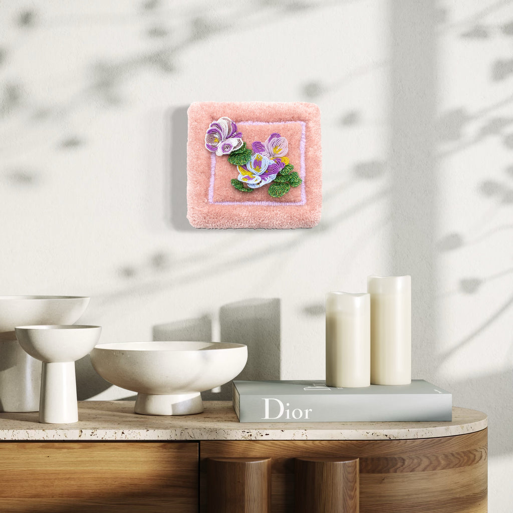 Pansies by Abigail Henthorne features a delicate textured artwork with soft purple and yellow floral motifs on a plush, muted pink square background, mounted on a light wall above a modern wooden sideboard displaying elegant white ceramic bowls, candles, and a hardcover book, creating a serene and refined atmosphere.