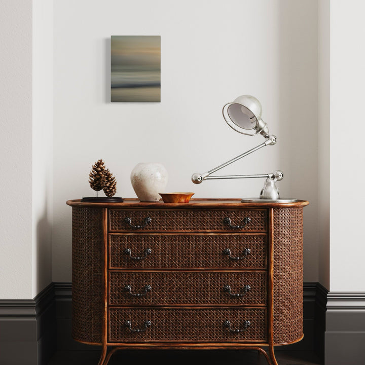 The artwork 'Sacred Cove' by Neil Shapiro features a serene abstract seascape print on textured Hahnemühle paper, displaying horizontal bands of muted earth tones blending from golden sunset hues to misty gray-blue horizons, creating a tranquil coastal atmosphere, mounted on a clean white wall above an antique dresser.