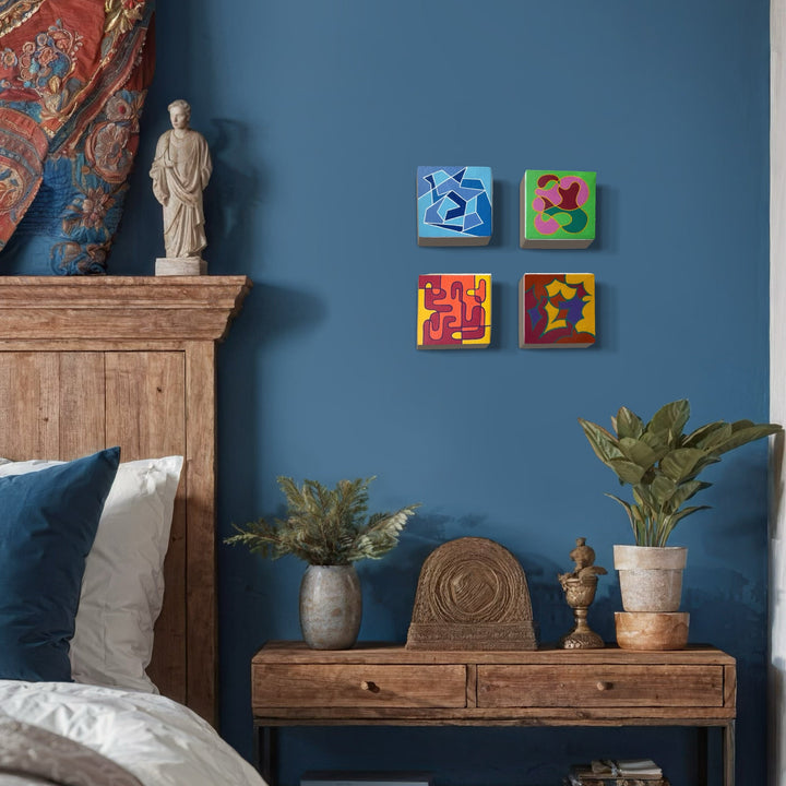 The artwork 'Seasonal Phenomena II' by Joe Piscopia features four vibrant square canvases arranged in a grid pattern, each featuring abstract geometric designs in bold primary colors against solid backgrounds. Blue, pink, yellow, and red compositions incorporate star and curved shapes, mounted on a deep blue wall in a bedroom setting with rustic wooden furniture and potted plants.