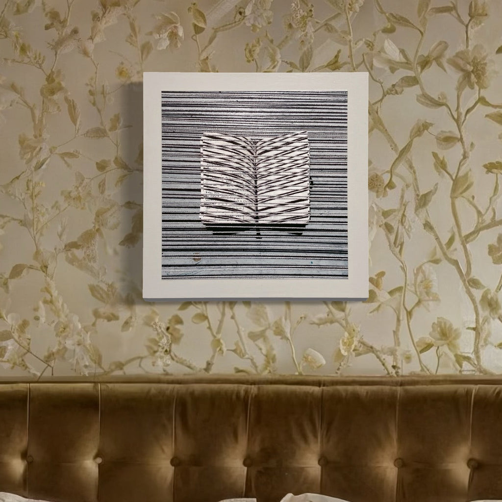 The artwork 'Sentence Structure String 6' by Anne Sherwood Pundyk features a striking monochromatic composition of parallel horizontal lines interrupted by a central white folded form resembling an open book or leaf pattern. The piece, mounted on a white panel against floral wallpaper, combines bold graphic elements with delicate textural details.