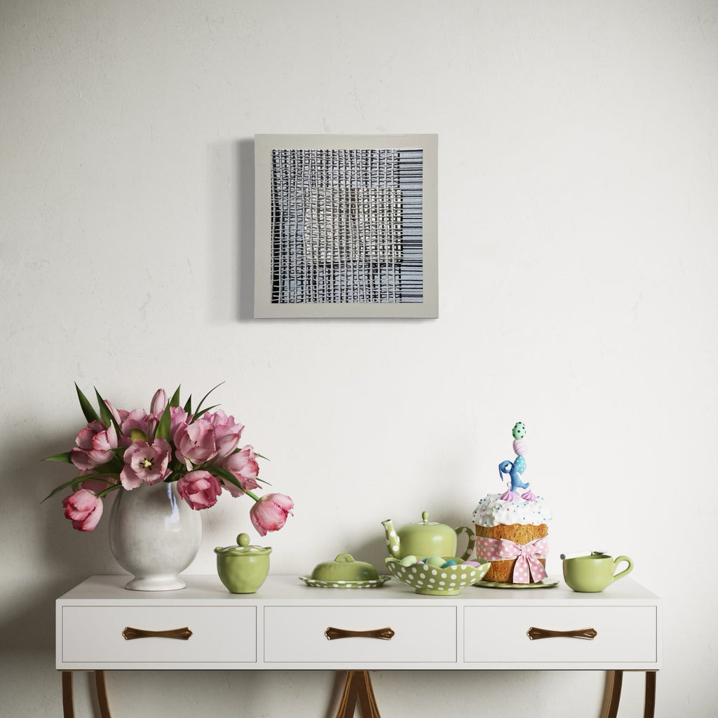 The artwork 'Sentence Structure String 7' by Anne Sherwood Pundyk hangs above a white console table featuring pink tulips in a ceramic vase, lime green tea set, and a decorated cake. The artwork displays a geometric black and white grid pattern with vertical and horizontal lines on a white panel, creating an abstract barcode-like composition. By Anne Sherwood Pundyk.