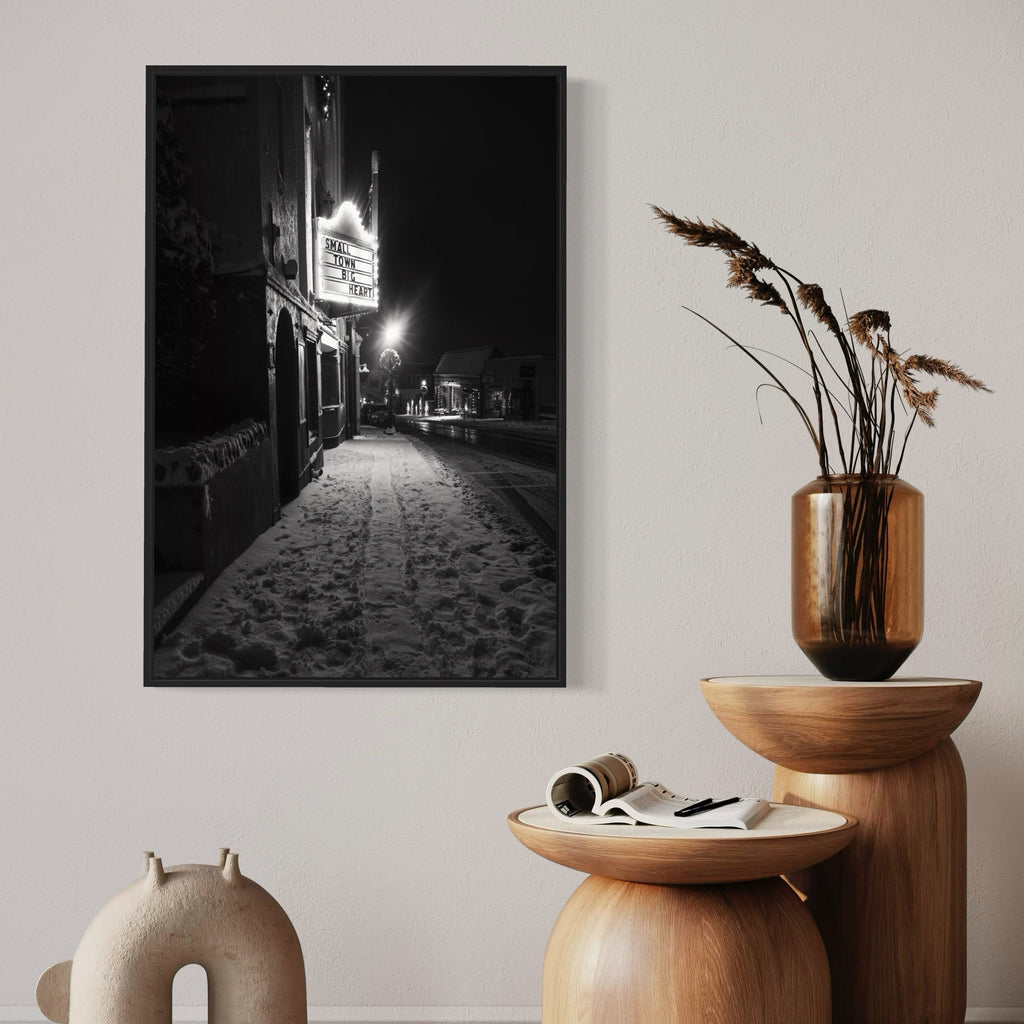 Small Town, Big Heart by Dennis Maida: Black and white nighttime photograph capturing a quaint small-town street scene. Illuminated storefront sign casts a warm glow on snow-covered sidewalk, creating dramatic contrast. Empty street stretches into darkness, bordered by historic buildings. Moody atmospheric composition in 36x24 format.