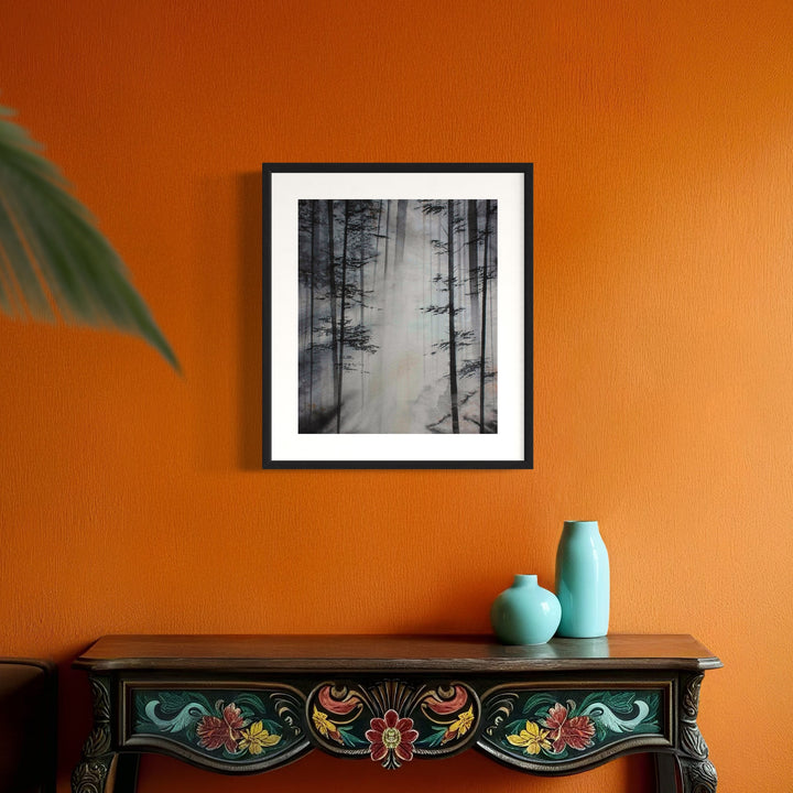 The artwork 'Songs of the Woodland' by Cat Lencke features a misty black and white forest scene with tall, silhouetted pine trees creating vertical lines against a foggy background. The atmospheric watercolor captures the ethereal nature of a woodland in winter, framed in classic black. Displayed against vibrant orange wall above decorative vintage furniture.