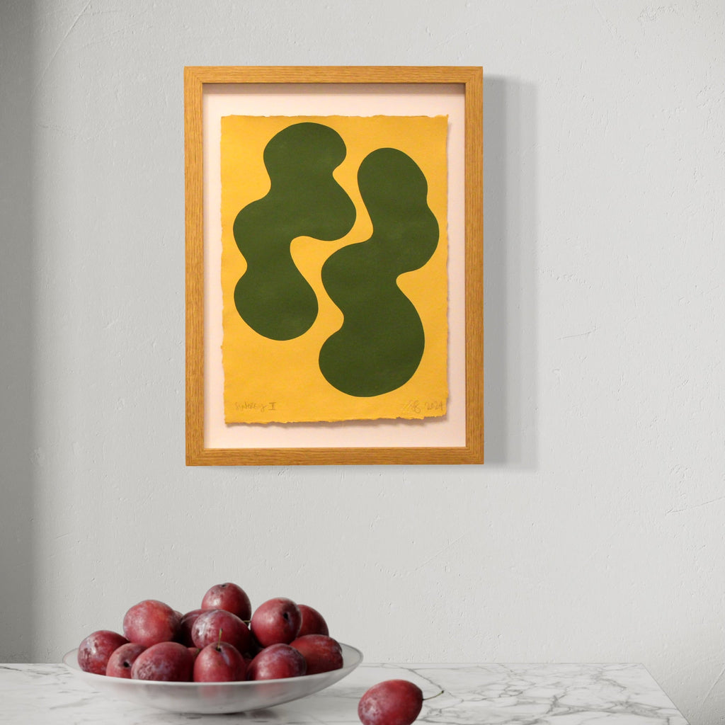 The artwork 'Synergy II' by Shadia Sabagh features two organic, flowing dark green shapes set against a vibrant yellow background, creating a bold minimalist composition. The abstract forms appear to dance and interact on handmade cotton paper, framed in natural wood. Acrylic painting with deckle edges showcases modern contemporary style.