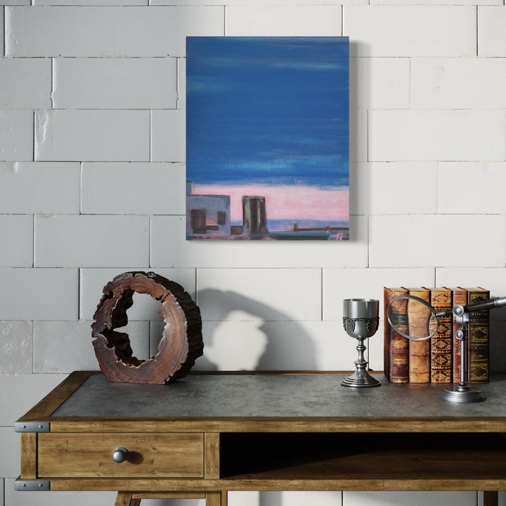 The artwork 'The Blue Hour (metallic)' by Cora Jane Glasser features an abstract cityscape in oil on linen, depicting architectural silhouettes against a deep blue twilight sky and soft pink horizon. Dark geometric shapes suggest buildings, while metallic undertones create a luminous dusk atmosphere. The 20x16 inch composition captures the transitional moment between day and night.