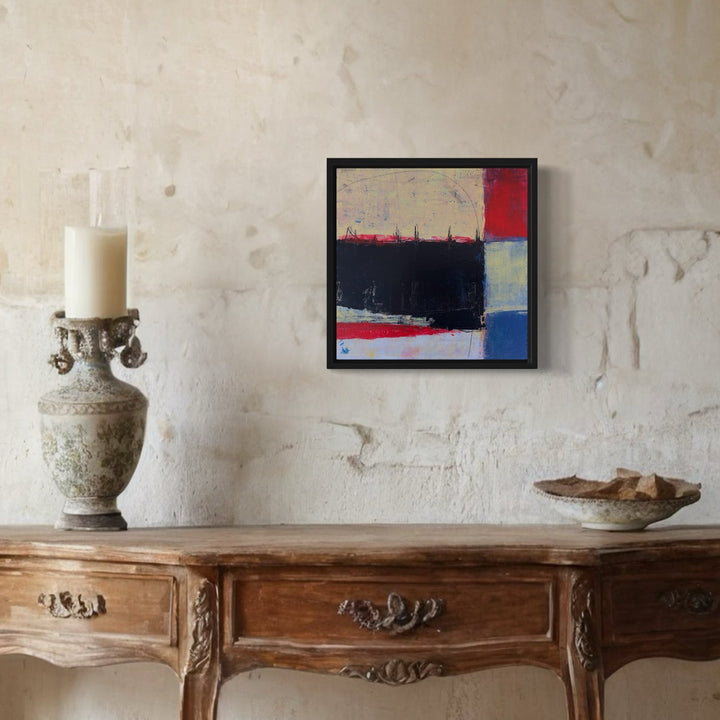 The artwork 'The Shield of Self' by Karen L. Burns features bold abstract composition with dramatic contrasts of black, red, and blue blocks against a textured backdrop. The acrylic painting showcases geometric forms with rough, expressive brushstrokes, mounted in a sleek black frame, creating a powerful contemporary statement piece that sits harmoniously above an antique wooden console.