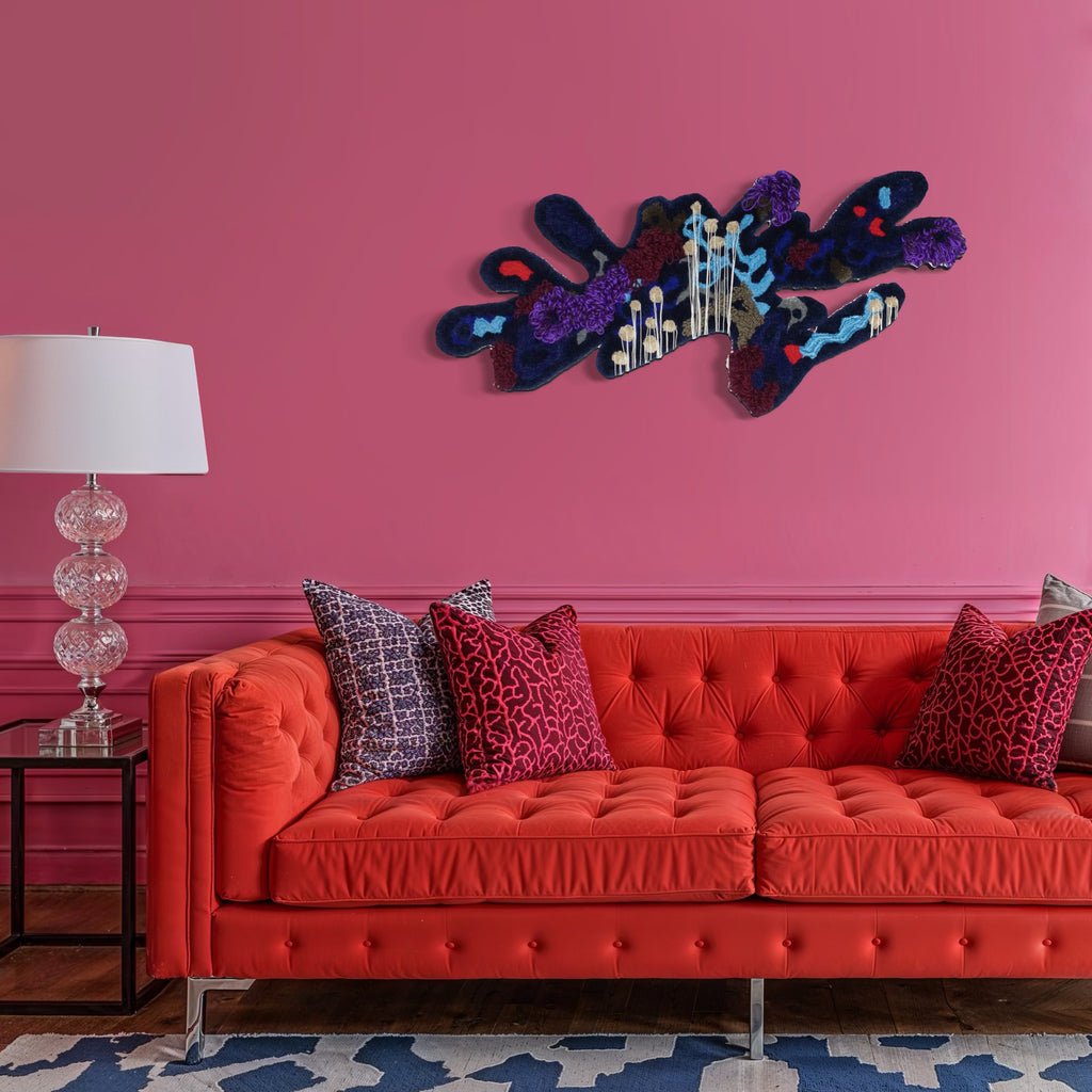 Vulturine Guinea Fowl Hen by Abigail Henthorne is a plush, abstract wall art piece featuring deep blues, purples, and soft cream tones with intricate texture and organic forms, set against a vibrant pink wall above a bold orange tufted sofa paired with patterned cushions and a glass table lamp, creating a lively yet sophisticated interior statement.