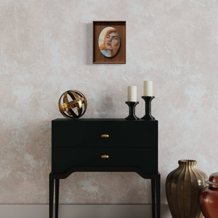 The artwork 'Dolly' by Jillian Shea showcases an oil painting mounted in a wooden frame, displayed above a black console table. The small-scale artwork features warm and cool tones against a textured background. Accompanied by decorative elements including brass spherical sculpture, white candles in black holders, and ceramic vases. 9.5x7.5 inches oil on canvas.