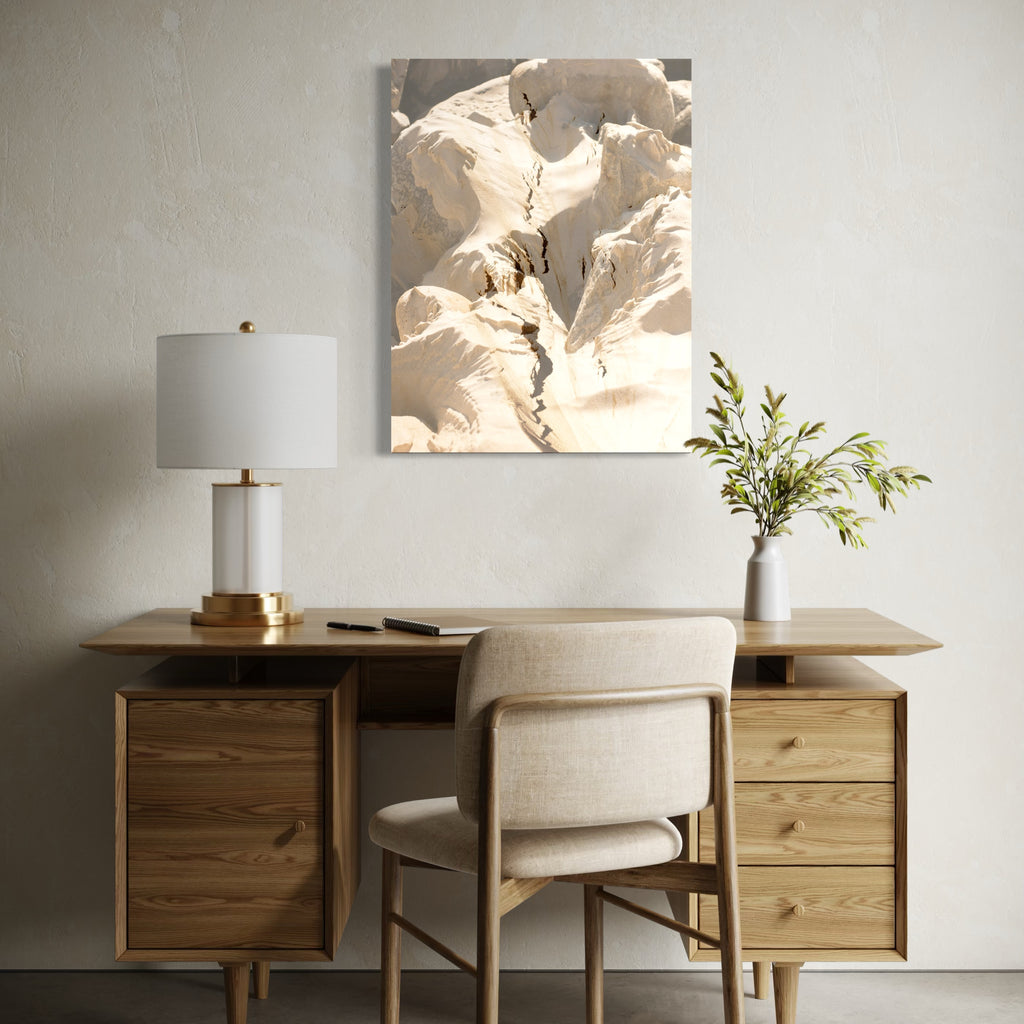 erosion by Eunice Pais depicts a close-up of cracked, sunlit earth with soft shadows highlighting textures and natural forms. Mounted above a minimalist wooden desk with muted decor and a vase of greenery, the artwork introduces a calm, organic feel that harmonizes with the serene, neutral-toned workspace.