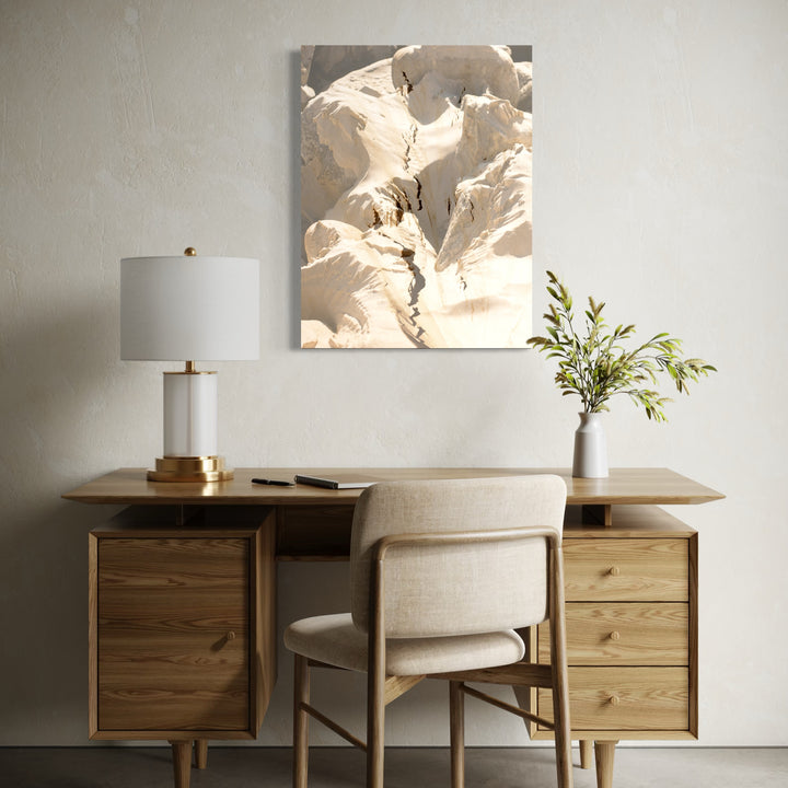 erosion by Eunice Pais depicts a close-up of cracked, sunlit earth with soft shadows highlighting textures and natural forms. Mounted above a minimalist wooden desk with muted decor and a vase of greenery, the artwork introduces a calm, organic feel that harmonizes with the serene, neutral-toned workspace.