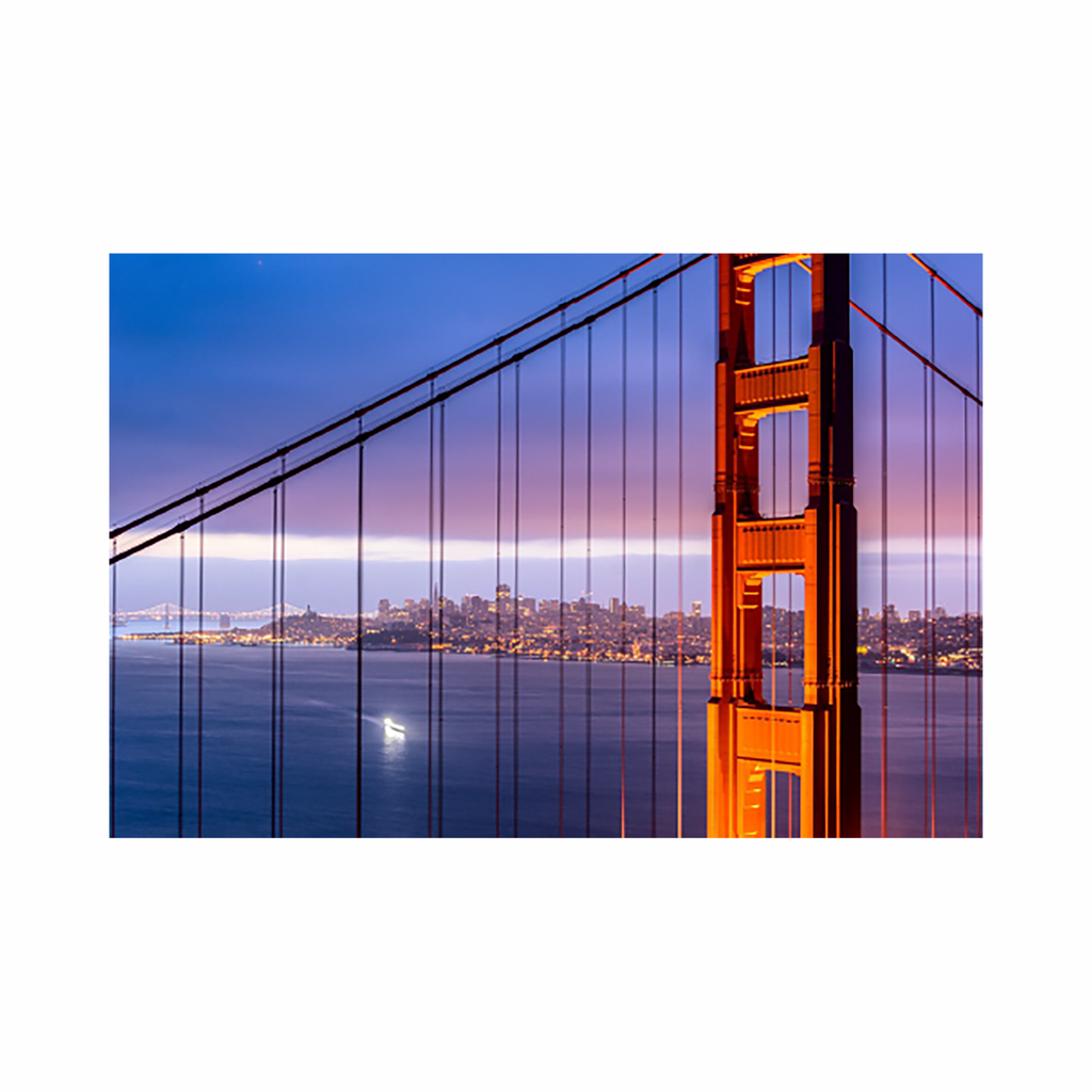 The artwork 'Suspension' by Dan Goldberger captures a close-up view of the Golden Gate Bridge's iconic orange tower against a purple twilight sky. The vertical suspension cables create strong leading lines, while the San Francisco cityscape glimmers in the background across calm blue waters, with a single boat's light reflecting on the bay surface.
