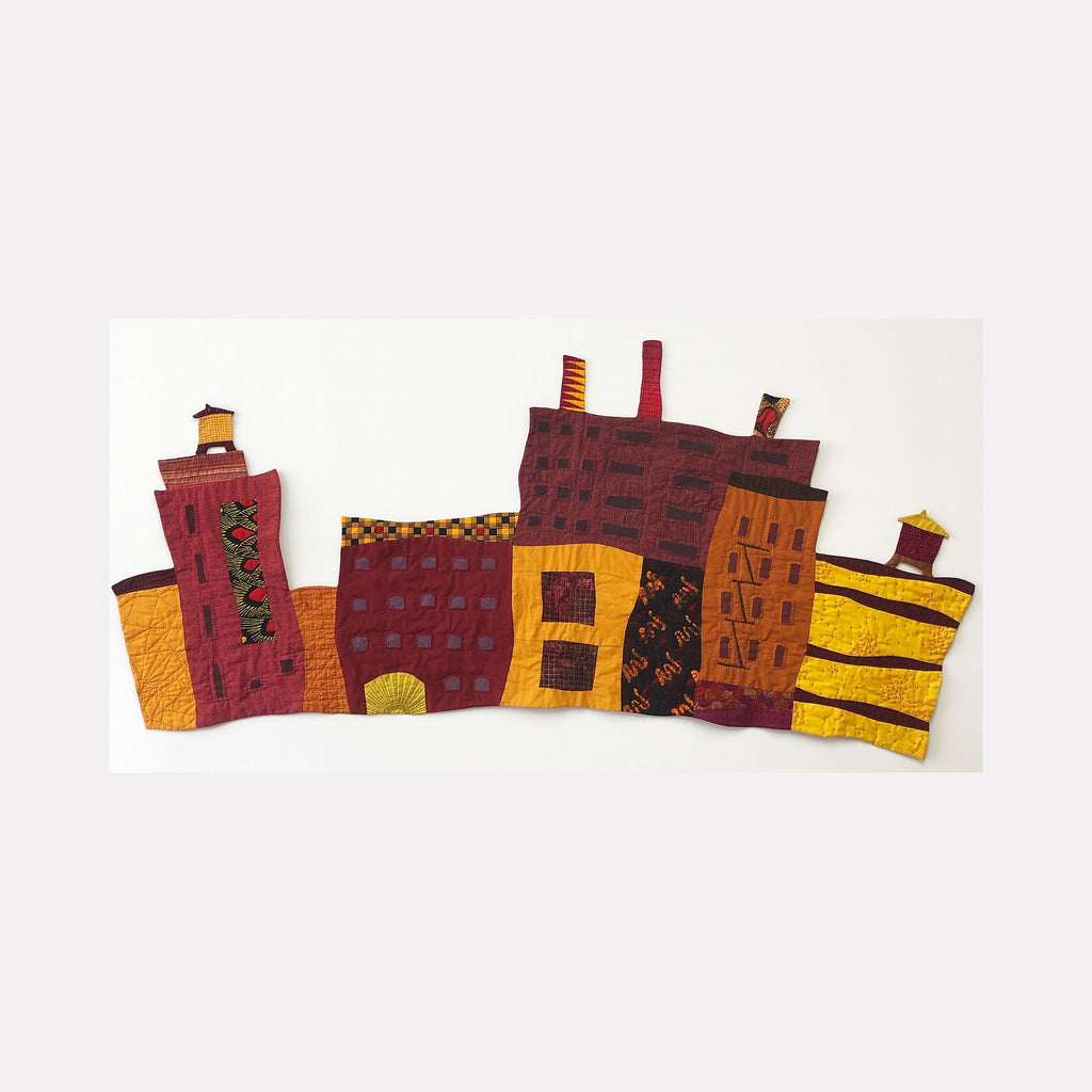 The artwork 'Cityscape with Smokestacks and Two Towers' by Ann Cofta features a vibrant textile collage depicting industrial buildings in burgundy and golden yellow. Hand-quilted fabric buildings with geometric patterns show smokestacks and architectural details, creating an abstract urban landscape against a white background. The mixed media piece combines applique work with paint detailing.