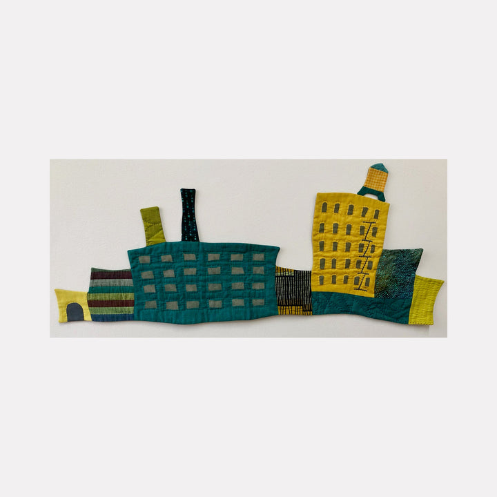 The artwork 'Cityscape (Shades of Teal & Mustard)' by Ann Cofta features an abstract urban skyline rendered in textile art, with geometric buildings in teal and mustard yellow. Hand-quilted fabric art depicts industrial structures with textured patterns, including factory-like buildings with chimneys and architectural details. Mixed media textile artwork showcases modern minimalist design.