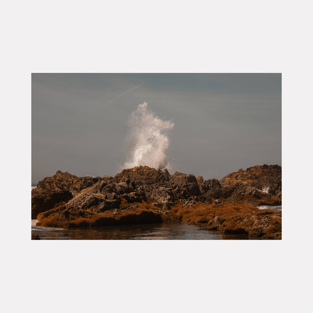 The artwork 'Communion V' by Eunice Pais captures a dramatic ocean wave crashing against rugged brown coastal rocks, creating a white spray explosion against a muted gray sky. The fine art photograph contrasts the ephemeral nature of water spray with the solid permanence of the weathered rocky shoreline in warm earthy tones. By Eunice Pais.