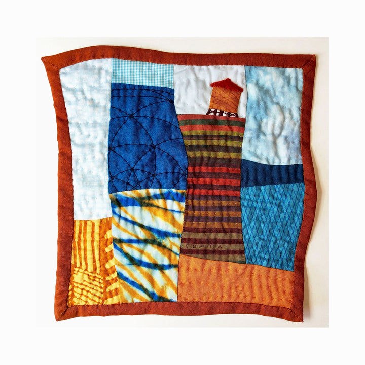 The artwork 'Cityscape (Shades of Blue & Orange)' by Ann Cofta features an abstract urban landscape crafted in quilted fabric. The composition combines geometric shapes in vibrant blues and warm oranges, with a prominent striped tower structure against a patchwork sky. Hand-quilted details and varied textile patterns create depth and texture in this 10x10 inch fiber art piece. Created by Ann Cofta.