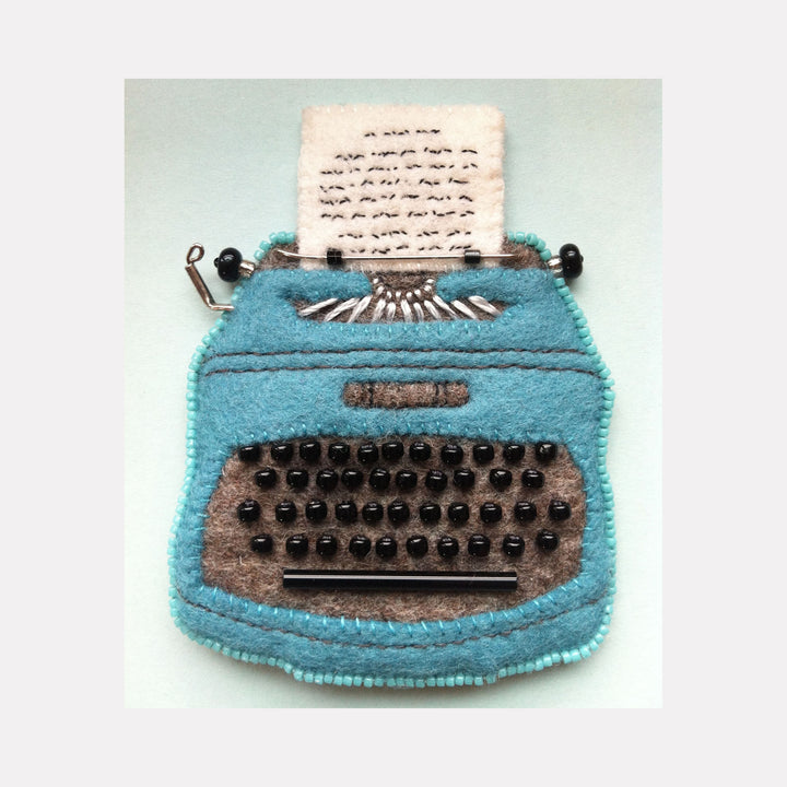 Vintage Typewriter (Olivetti Lettera) by Ann Cofta: Handcrafted felt artwork featuring a turquoise typewriter with gray keyboard, detailed with black beaded keys, wire accents, and hand embroidery. Paper sheet emerges from the platen, displayed in shadowbox frame against mint background.