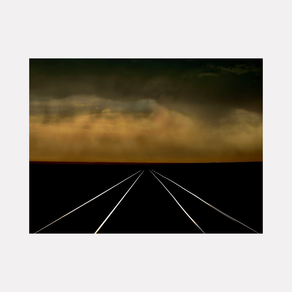 The artwork 'Bakersfield Tracks' by Neil Shapiro captures dramatic railway lines converging to a distant vanishing point against a dark foreground, set beneath an atmospheric golden-amber sky with moody storm clouds. The stark geometric lines of the tracks create powerful perspective on textured Hahnemühle print medium.