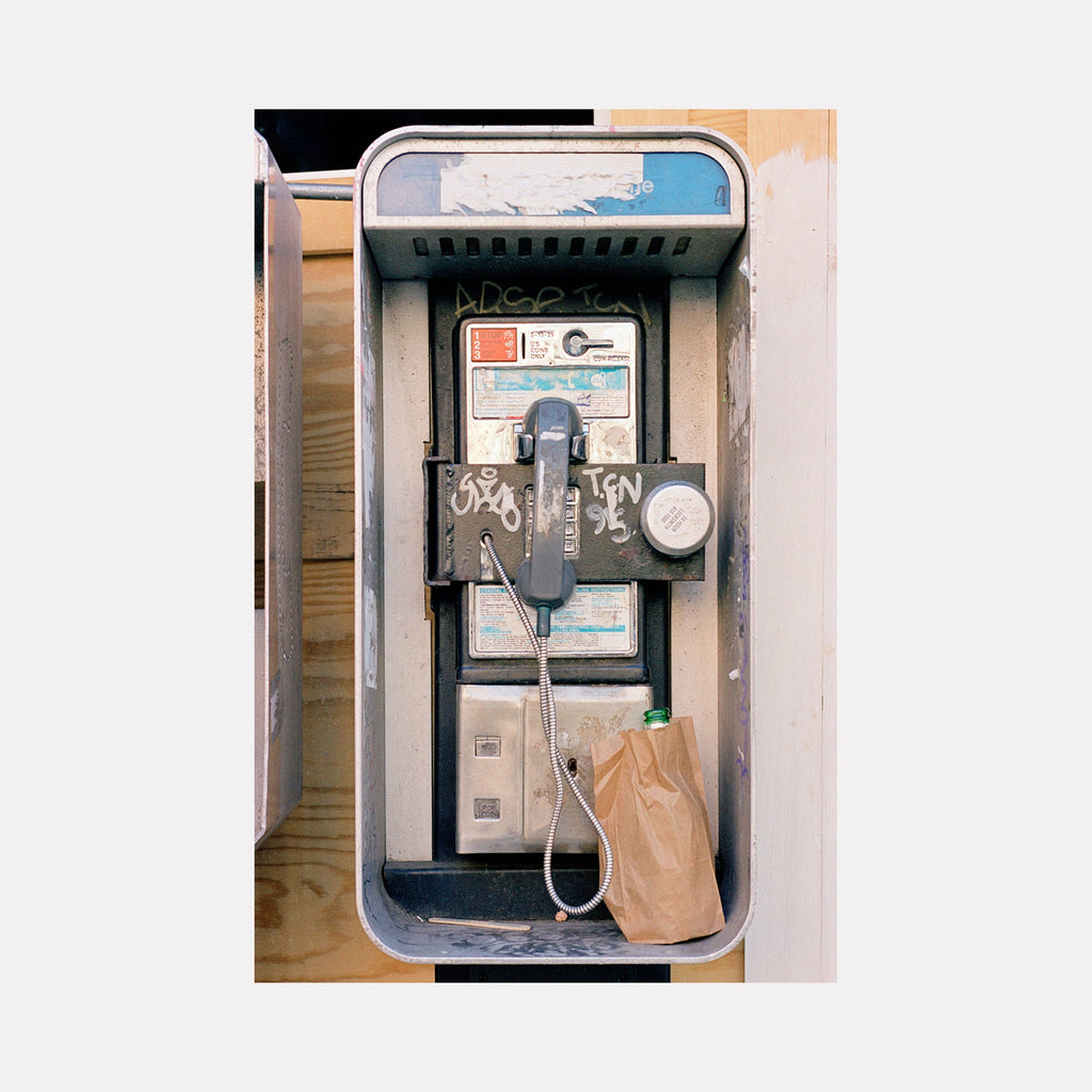 The artwork 'Brown Bag' by Dirk Westphal captures a vintage payphone booth with a brown paper bag, featuring an urban nostalgic composition. The payphone shows weathered graffiti marks against a metallic enclosure, with a curved top frame and blue-tinted background. The editioned signed photograph measures 30x20 inches, presenting an authentic slice of city infrastructure.