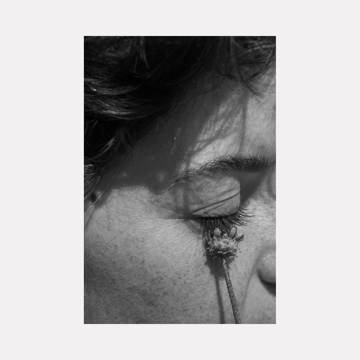 The artwork 'Communion II' by Eunice Pais presents a striking black and white photography composition showing a close-up of a closed eye with a delicate twig creating a gentle shadow against skin texture. The intimate monochromatic detail captures subtle contrasts and emotional depth through sharp focus and dramatic lighting. By Eunice Pais.