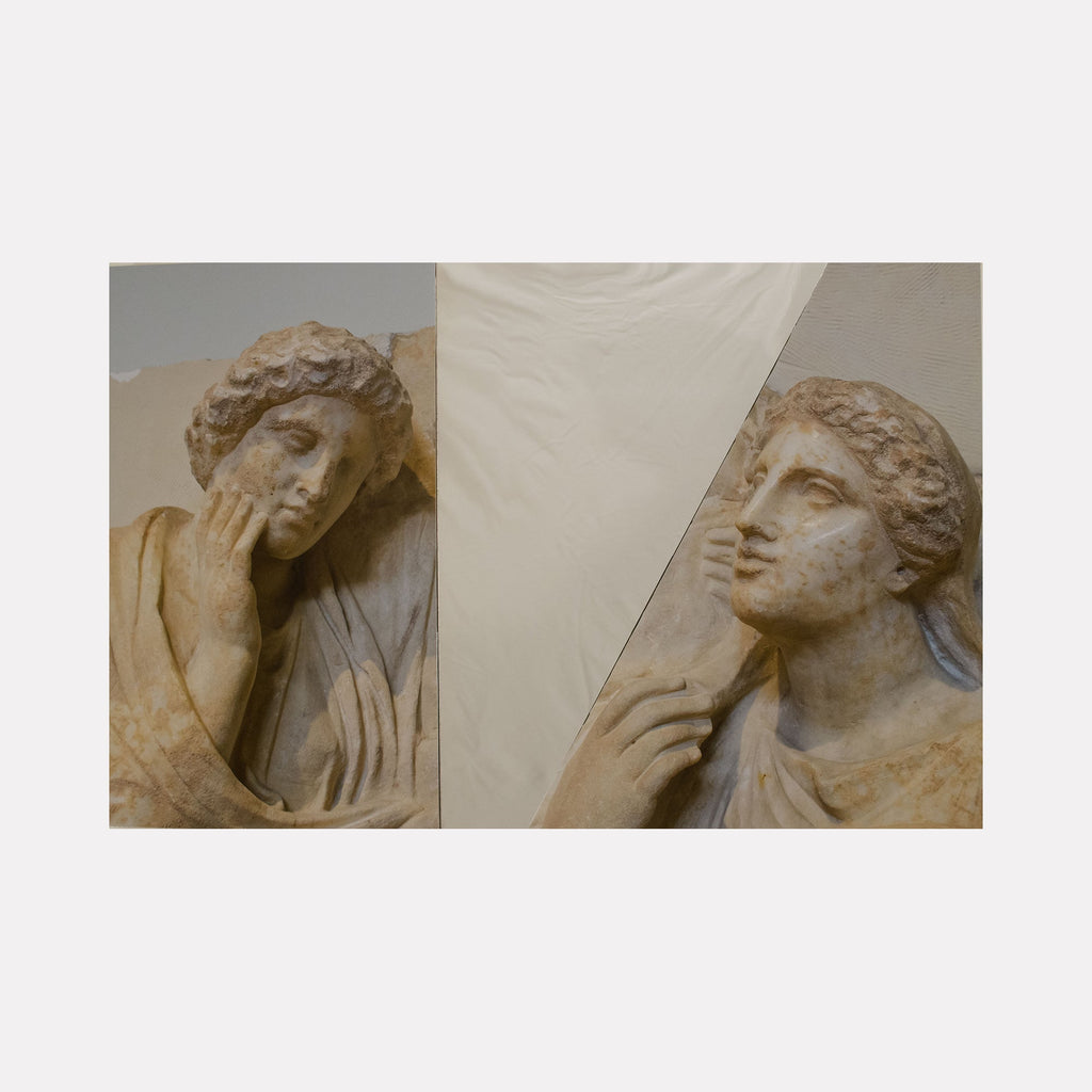 The artwork 'Grave Mirror' by Ryan Frank features a split composition of classical Greek-style sculpture reliefs captured in a photograph. A mirrored diptych shows weathered stone figures with curled hair and draped garments in soft beige tones, each figure making contemplative gestures. The artwork combines photography and mirror elements in a wooden frame.