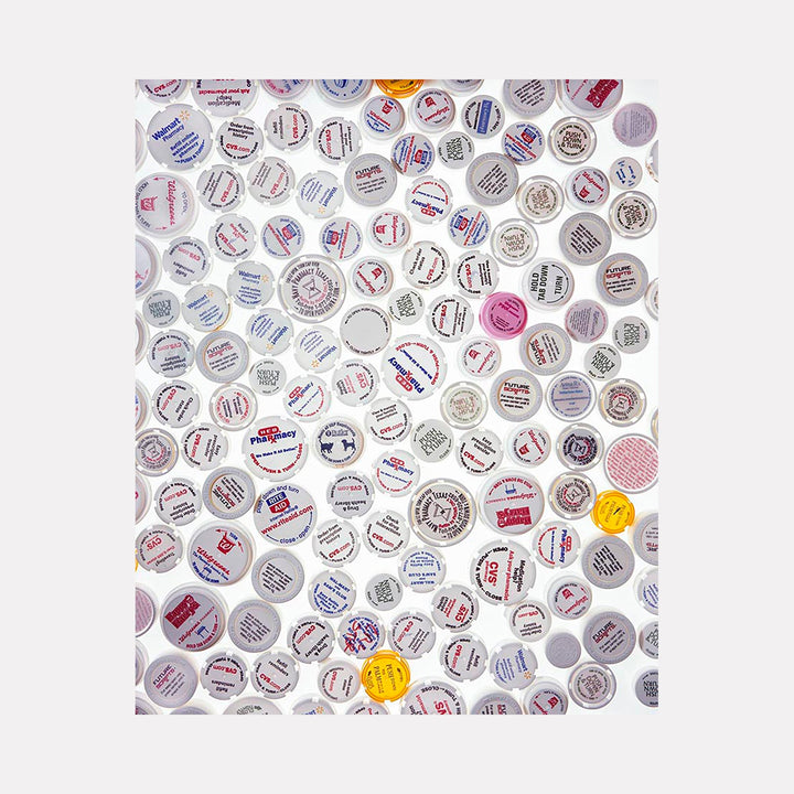 The artwork 'RX' by Dirk Westphal features a mesmerizing collage of circular prescription bottle caps arranged in a scattered pattern against a white background. The caps display various pharmaceutical text and labels in red, blue, and gray tones, with occasional pops of yellow, creating a contemporary commentary on medication culture. Signed editioned photograph by Dirk Westphal.