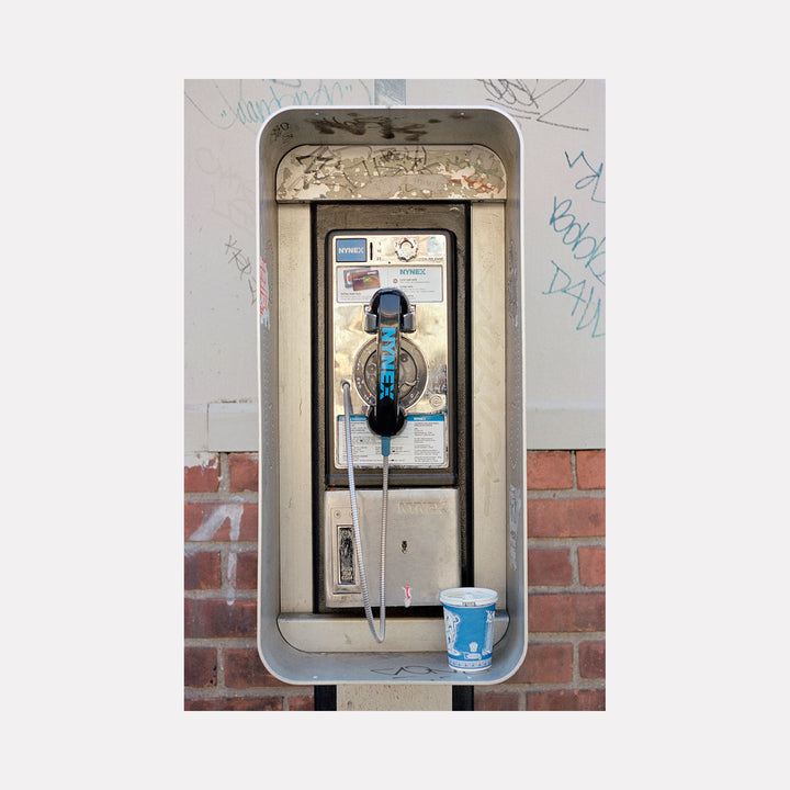 The artwork 'Rotary:Coffee' by Dirk Westphal captures a vintage payphone installation with a disposable coffee cup placed within its metal enclosure. The weathered white payphone features blue accents and silver metallic details, mounted on a brick wall with graffiti marks. Editioned signed photograph, 30x20 inches by Dirk Westphal.