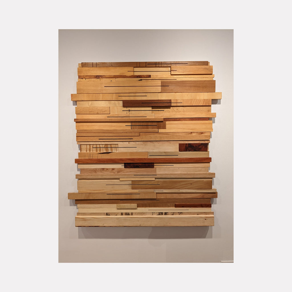 The artwork 'These Woods Were Trees II' by Ryan Frank features a striking wall sculpture composed of layered wooden strips arranged horizontally, creating a dynamic textural composition. Various wood types including birch, mahogany, and cedar create a natural gradient of honey, amber, and golden tones. The pieces are mounted to form a contemporary abstract pattern with varying depths and shadows.