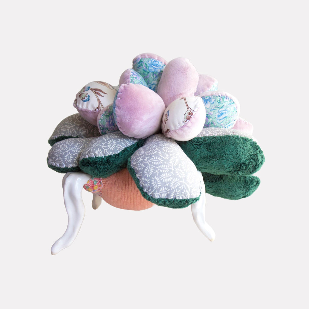 Four-Legged Plume by Hanna Washburn is a tactile textile sculpture featuring plump, cushioned sections in soft pinks, patterned blues, and deep green, arranged like oversized petals atop elegant, curved white legs. The piece blends fabric textures and whimsical forms to create an inviting, imaginative art object with a playful, organic presence.