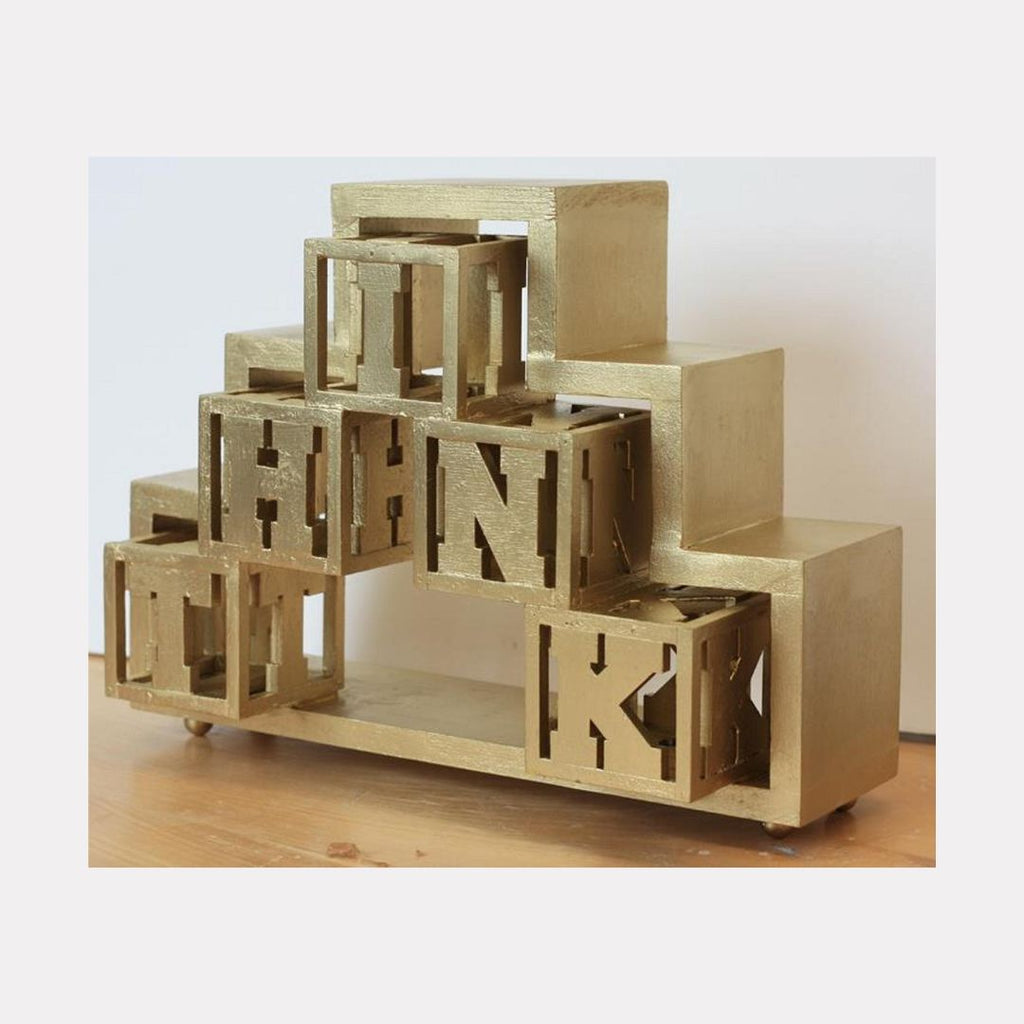 The artwork "Think Gold" by Carl Chaiet in gallery position 1 presents a sculptural arrangement of gilded cubes and rectangular blocks, each containing cut-out letters in a bold geometric style. The metallic gold finish and structured composition recall the minimalist abstraction of Donald Judd and the architectural elegance found in works by Sol LeWitt.