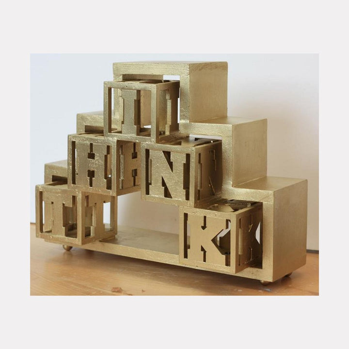 The artwork "Think Gold" by Carl Chaiet in gallery position 1 presents a sculptural arrangement of gilded cubes and rectangular blocks, each containing cut-out letters in a bold geometric style. The metallic gold finish and structured composition recall the minimalist abstraction of Donald Judd and the architectural elegance found in works by Sol LeWitt.