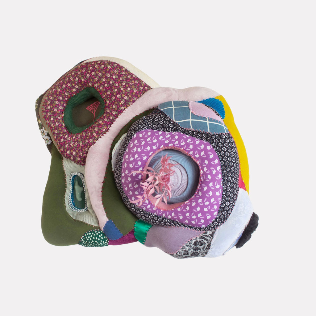 Wallflower by Hanna Washburn is a layered textile sculpture featuring concentric organic shapes in varied fabrics including floral prints, geometric patterns, and textured solids in colors like purple, green, pink, and yellow. The piece combines intricate stitching and plush textures to evoke a sense of softness and depth in an abstract, tactile form.